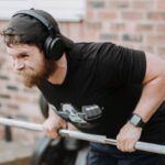 Brick Workouts - Brutal male with headphones pulling massive metal barbell on blurred background of brick house