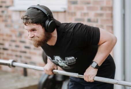 Brick Workouts - Brutal male with headphones pulling massive metal barbell on blurred background of brick house