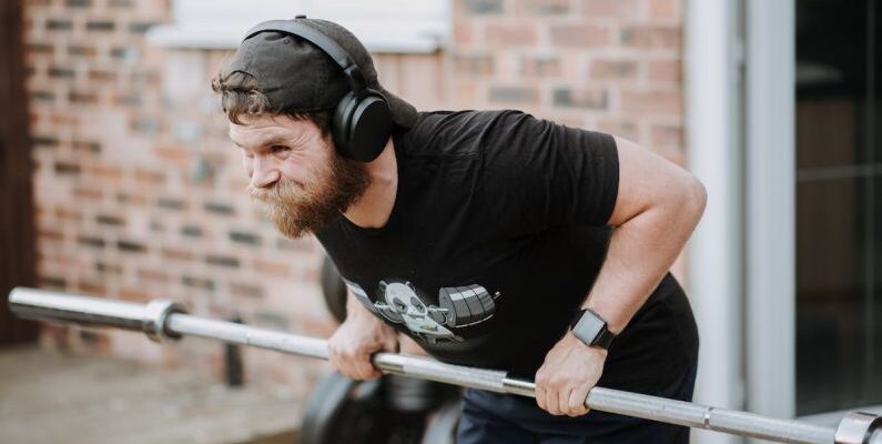 Brick Workouts - Brutal male with headphones pulling massive metal barbell on blurred background of brick house