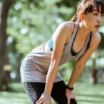 Recovery - Tired ethnic female in sportswear looking away while leaning on knees having rest after workout in park on sunny summer day