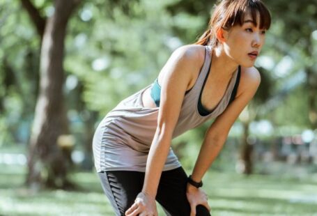 Recovery - Tired ethnic female in sportswear looking away while leaning on knees having rest after workout in park on sunny summer day