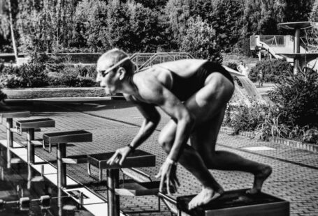 Swim Starts - Black and white side view full body sporty swimmer in swimming suit and goggles standing on block in track start position preparing to dive in outside pool