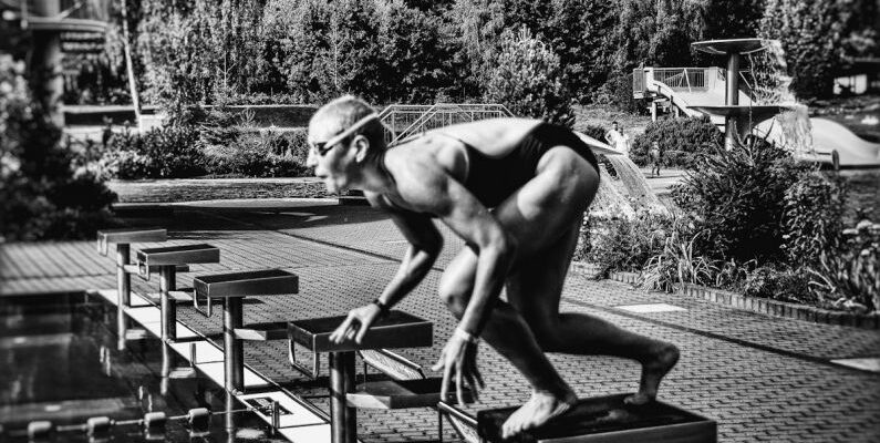 Swim Starts - Black and white side view full body sporty swimmer in swimming suit and goggles standing on block in track start position preparing to dive in outside pool