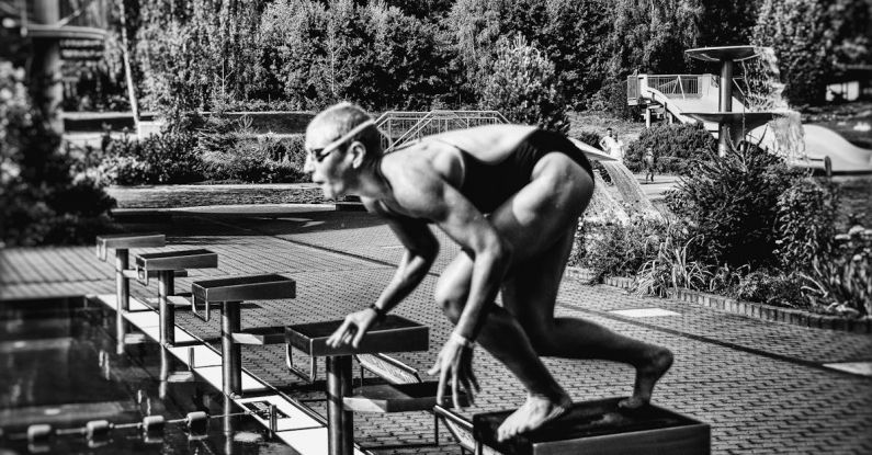 Swim Starts - Black and white side view full body sporty swimmer in swimming suit and goggles standing on block in track start position preparing to dive in outside pool
