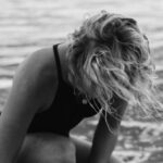 Water Games - Black and white of female surfer in swimwear sitting in foamy water of sea while fastening leash on leg