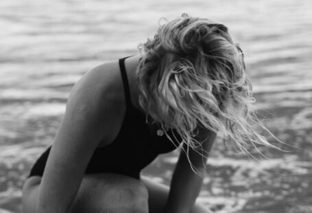 Water Games - Black and white of female surfer in swimwear sitting in foamy water of sea while fastening leash on leg