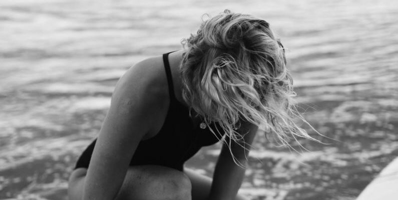 Water Games - Black and white of female surfer in swimwear sitting in foamy water of sea while fastening leash on leg