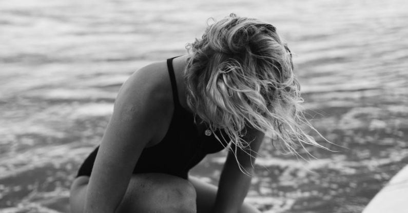 Water Games - Black and white of female surfer in swimwear sitting in foamy water of sea while fastening leash on leg