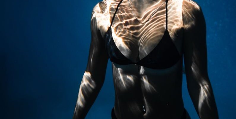 Dive Techniques - Underwater shot of fit young female in black bikini swimming in dark blue seawater with eyes closed